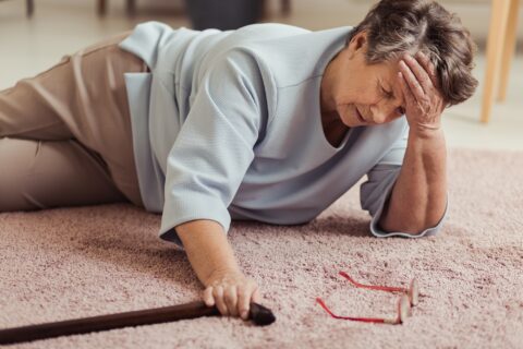 A woman who has fallen on the ground. She is holding her head in her hands as her cane and glasses lay on the floor beside her.